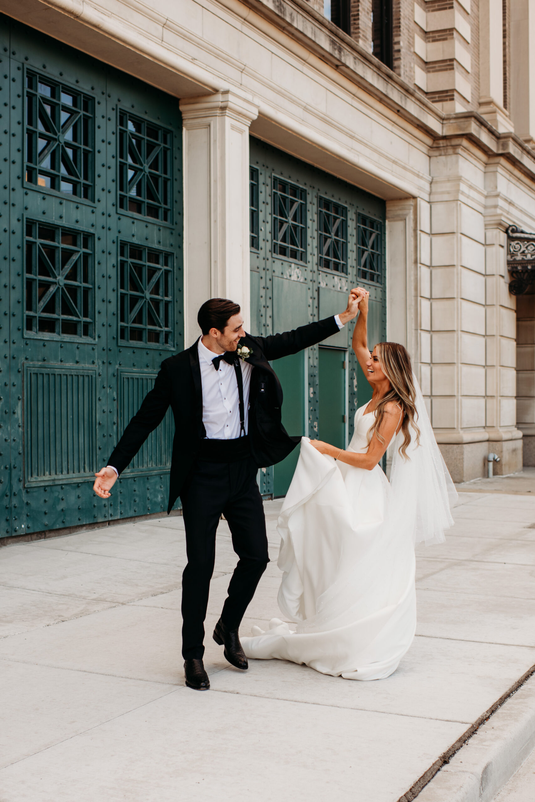 bride and groom in classic wedding attire dancing outside on the street in front of a blue wall near