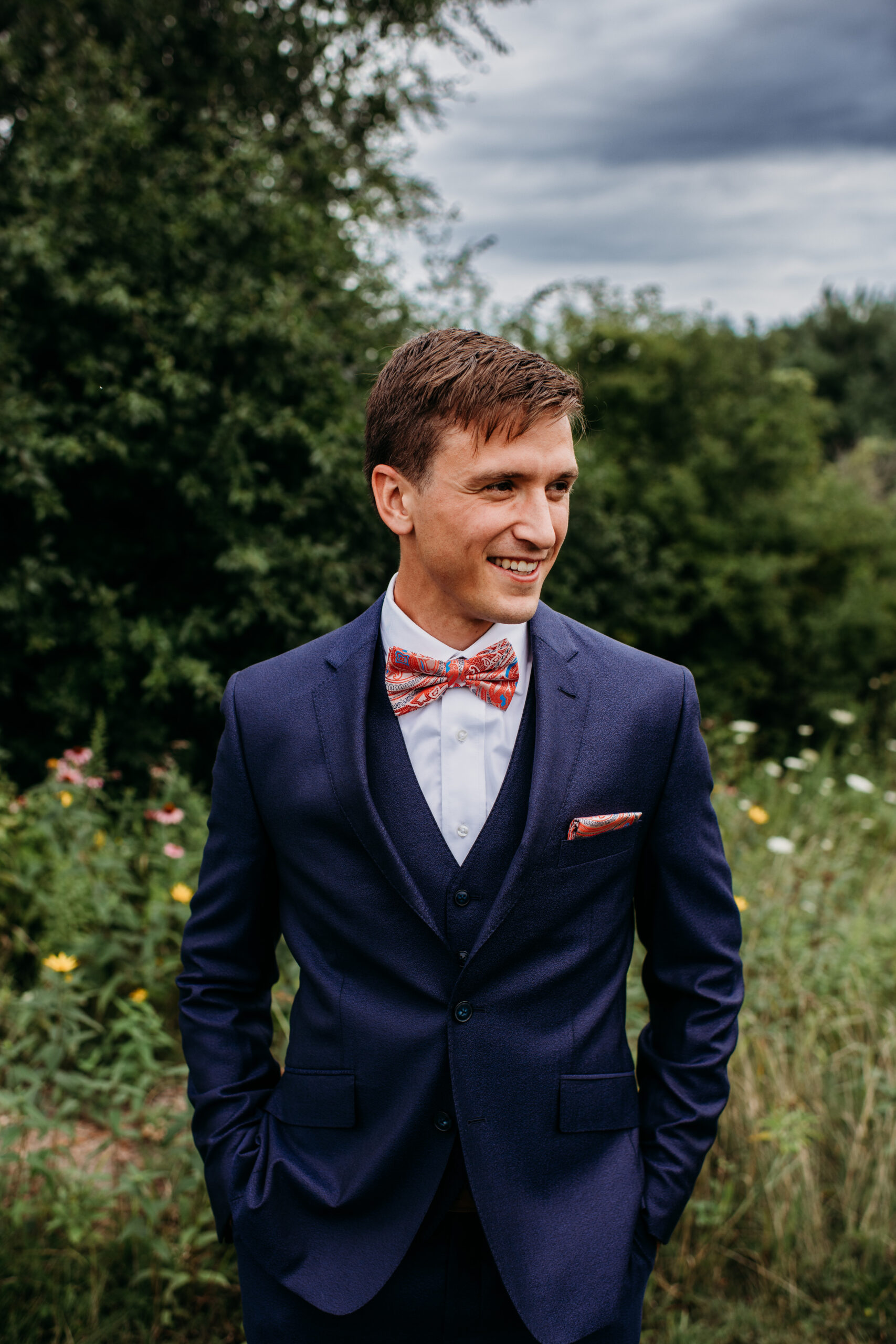 Groom in navy suit and salmon bowtie looking off into the distance in a field